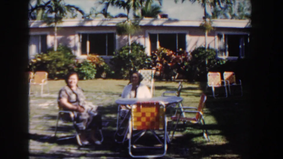 Two women sit at a round table in a Nassau garden, with palm trees, shrubs, woven chairs and a low house visible behind them.