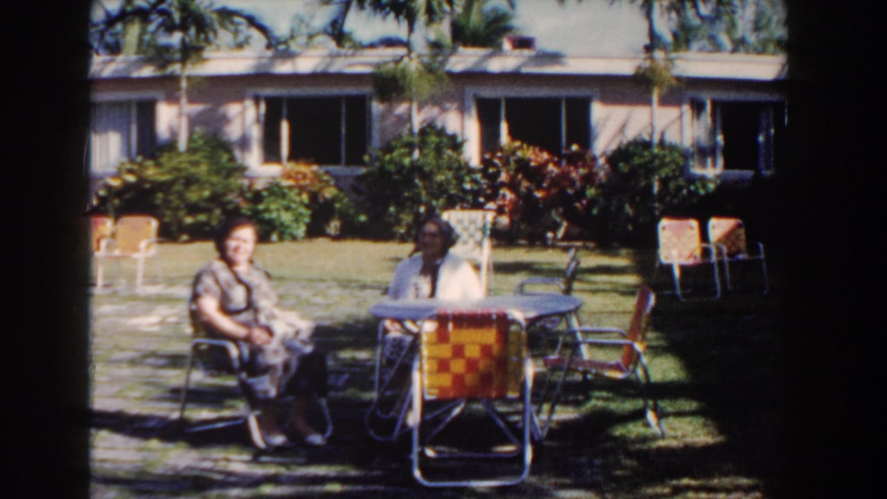 Two women seated outside beside orange chairs in a palm-shaded garden.