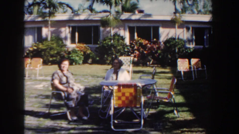 Two adult women sit on a lawn near woven patio chairs in Nassau, Bahamas, with palms and a pink stucco building behind them.
