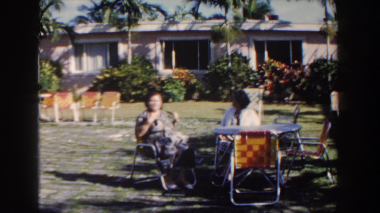Two women sit at a garden table with palm trees and a low building behind them.
