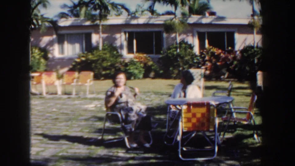 In a 1961 Nassau garden scene, two women sit near a round table and bright woven chairs under palm shade.