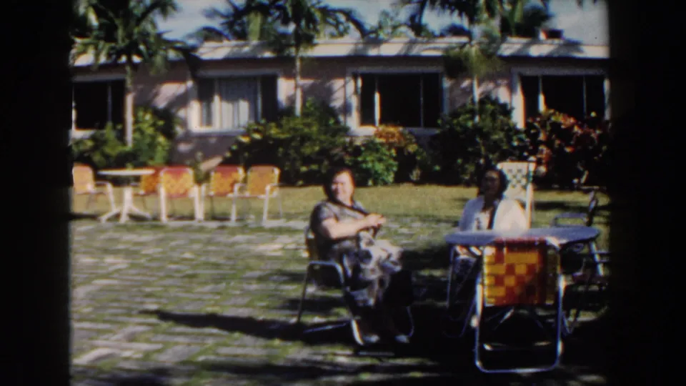 Two women sit in colorful garden chairs in Nassau, Bahamas, with a palm-lined lawn and stucco building in the background.