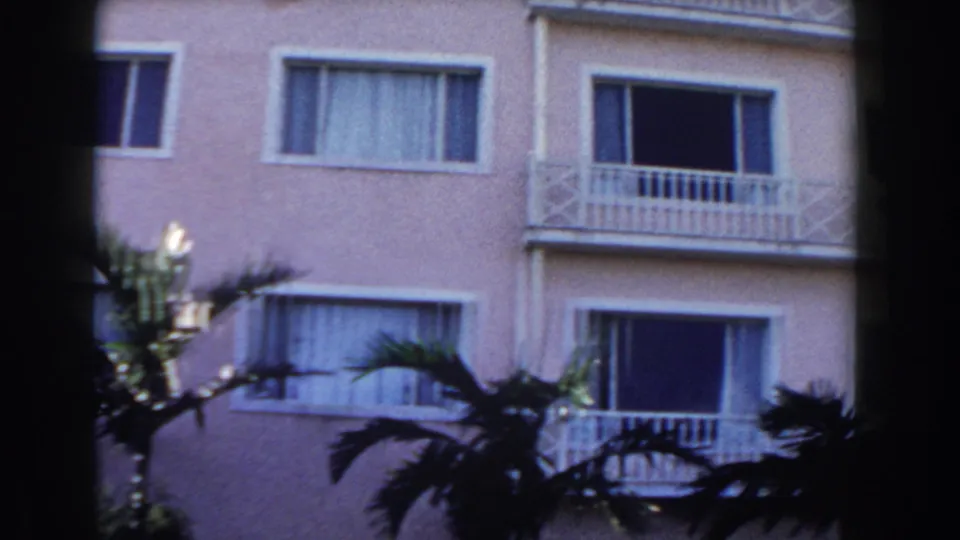 1961 Nassau home-movie frame showing a pale multi-level building with white balcony railings, dark windows, and palm fronds in front.