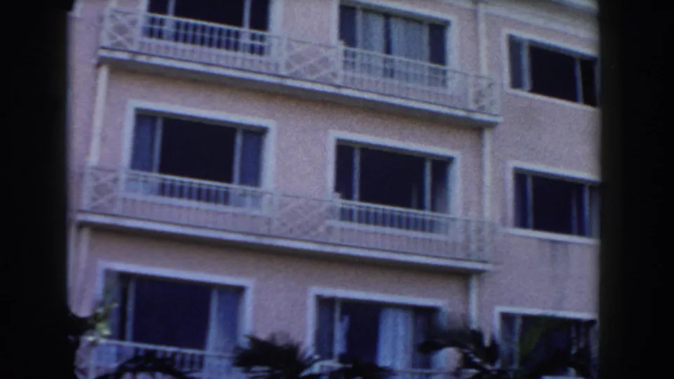 1961 Nassau frame of a pale building exterior with repeated windows, white balconies, and dark palm trunks crossing the view.