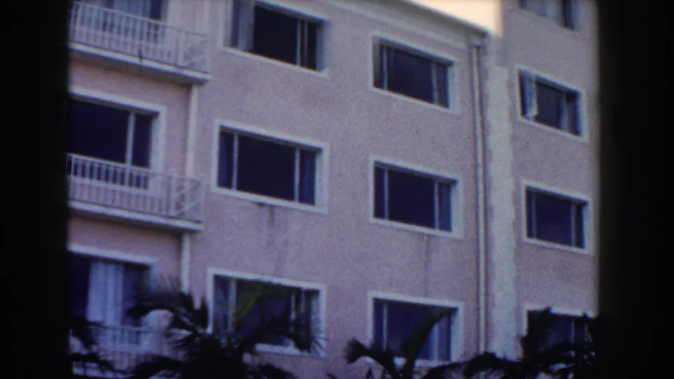 1961 Nassau home-movie frame showing the side of a pale multi-level building, rectangular windows, partial balconies, and palms below.