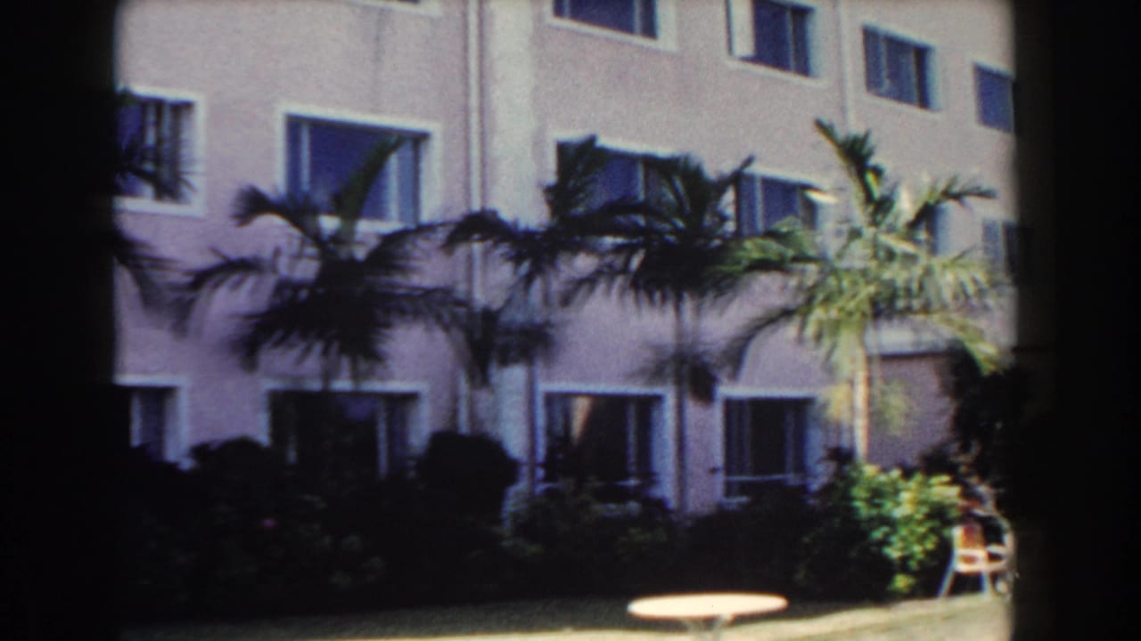 1961 Nassau hotel grounds with a pink facade, white-trimmed windows, palms, shrubs, and patio seating in soft 8mm color.