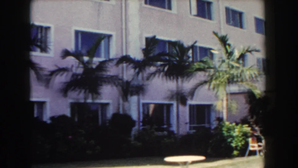 1961 Nassau hotel grounds with a pink facade, white-trimmed windows, palms, shrubs, and patio seating in soft 8mm color.