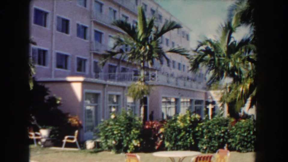Wider 1961 Nassau resort view with pink hotel walls, palms, hedges, balconies, a round table, chairs, and lawn.