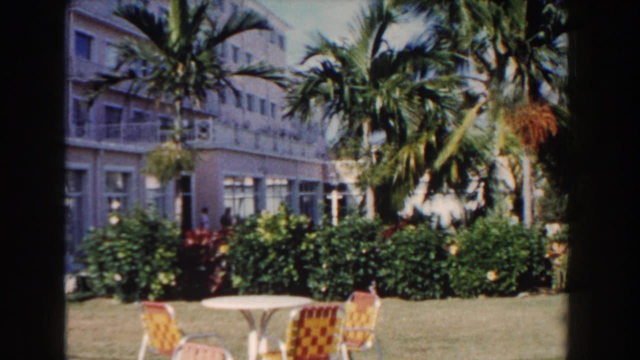 1961 Nassau resort grounds with palms, hedges, a hotel facade, white garden table, and patterned patio chairs on the lawn.
