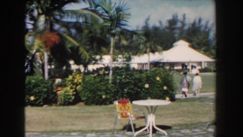 1961 Nassau resort garden with palms, white patio table, patterned chairs, trimmed hedges, and two guests walking near a low building.