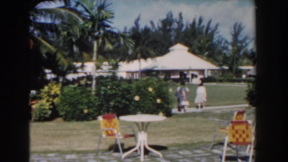 1961 Nassau resort grounds showing palms, hedges, a white table, patterned chairs, and distant guests walking beside low hotel buildings.