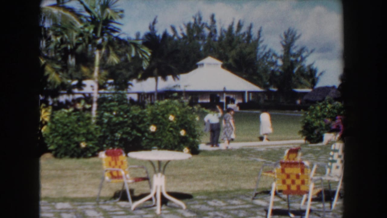 Resort lawn with patio chairs, flowering shrubs, and adults walking near a white-roofed building.
