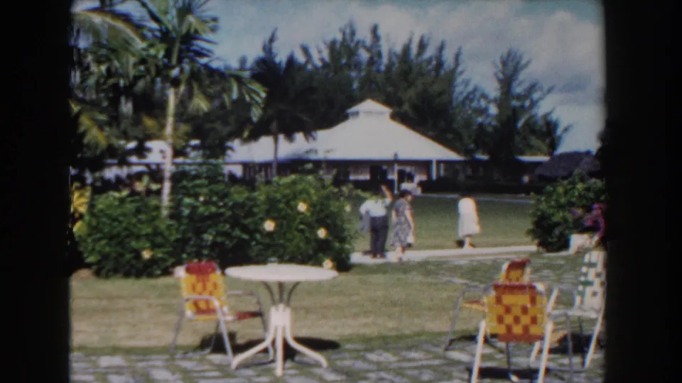 1961 Nassau resort grounds with woven chairs, a white table, palms, flowering bushes, and adults strolling on a sidewalk.