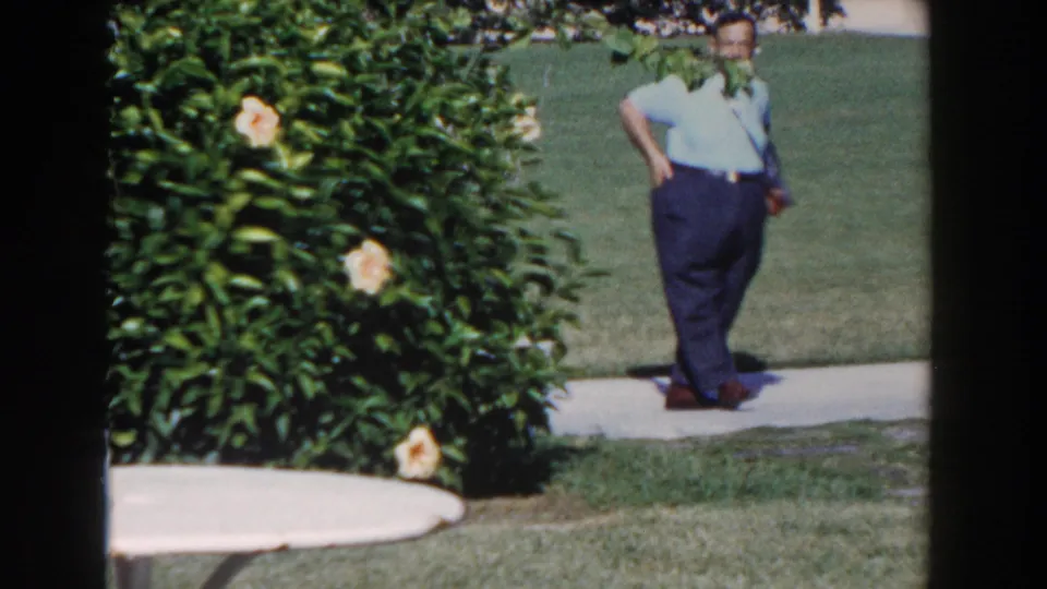 1961 Nassau resort frame showing a man near the walkway, with flowering shrubs, lawn, patio furniture, and soft 8mm color.