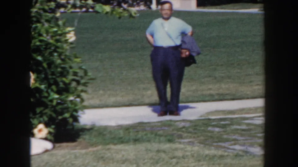 Nassau resort home-movie frame of a man standing on a garden sidewalk near lawn, flowering shrubs, and patio seating.