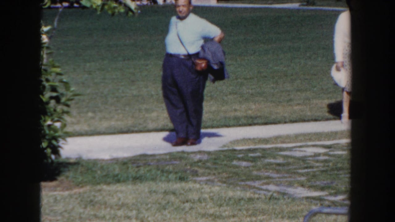 In Nassau, Bahamas, 1961, a man in a light blue shirt and dark trousers walks beside a lawn path with film frame edges visible.
