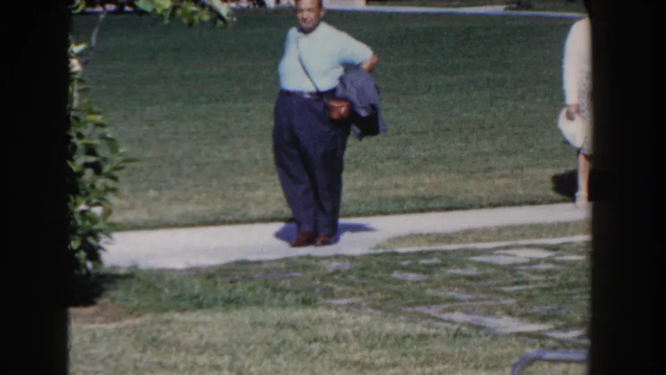 In Nassau, Bahamas, 1961, a man in a light blue shirt and dark trousers walks beside a lawn path with film frame edges visible.