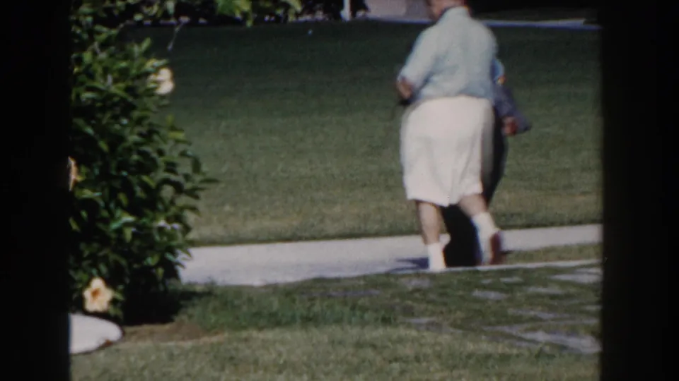 A woman walks away along the Nassau resort path, framed by grass, shrubs, and visible 8mm film gate edges.