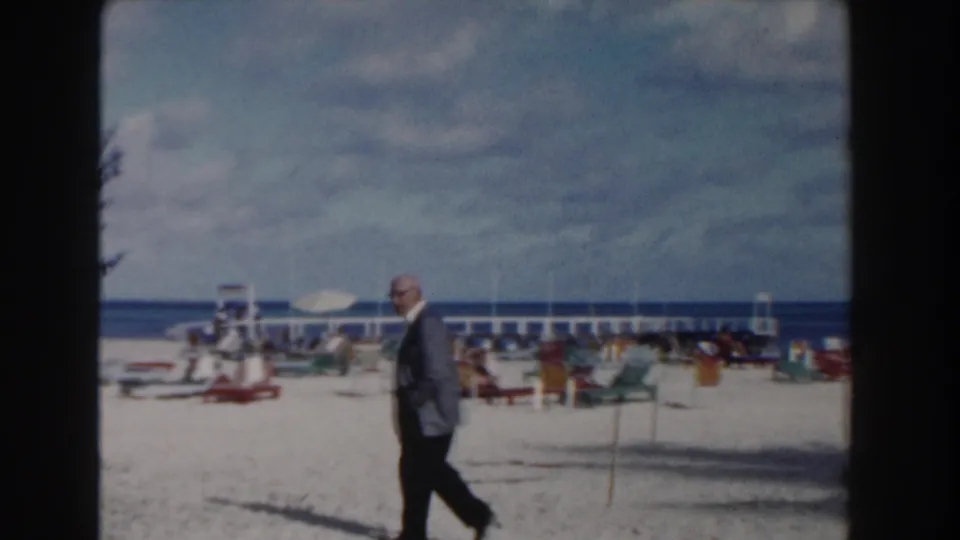A suited man walks across a Nassau beach in 1961, with lounge chairs, umbrellas, a rail or pier, sand, sea, and cloudy sky behind him.