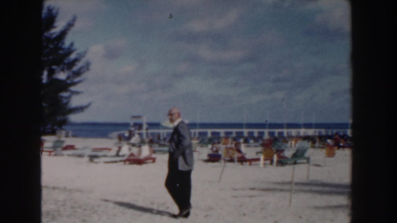 Man in a jacket walks across a bright beach with chairs and ocean behind him.