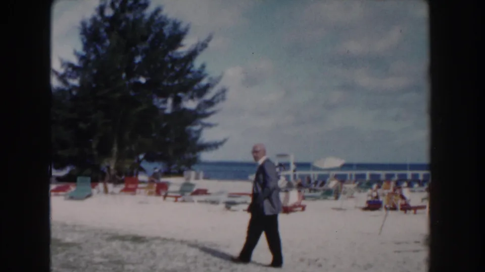 The man continues across the Nassau beach, passing lounge chairs beneath a large tree with the ocean visible beyond.