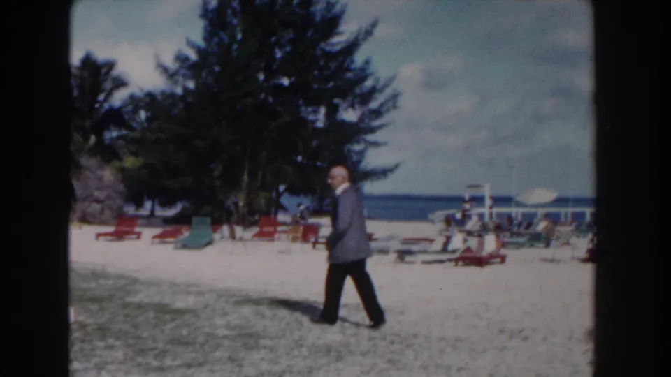 A 1961 Nassau beach view shows the man walking through the foreground while resort chairs, trees, umbrellas, and ocean sit behind him.