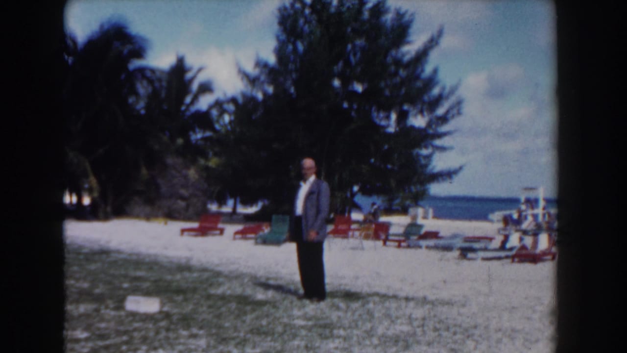 Man in a suit stands on a beach with lounge chairs, trees, and ocean behind him.