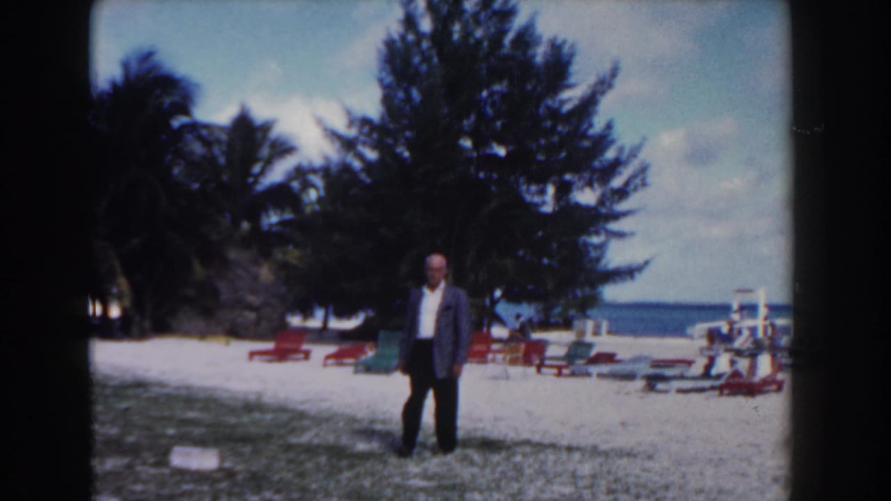 Man in a suit walks forward on a beach with colorful lounge chairs and trees behind him.