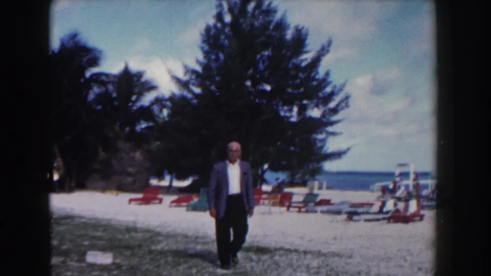 A suited man walks closer to the camera on the Nassau beach, with red lounge chairs, trees, sand, and ocean in view.