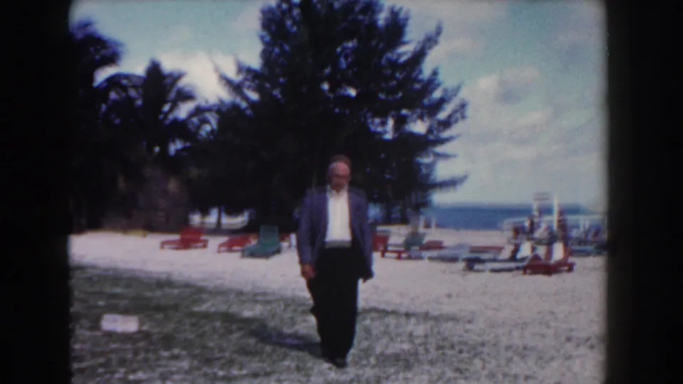 Near the end of the shot, the suited man is closer to the camera on the 1961 Nassau beach with chairs and water behind him.