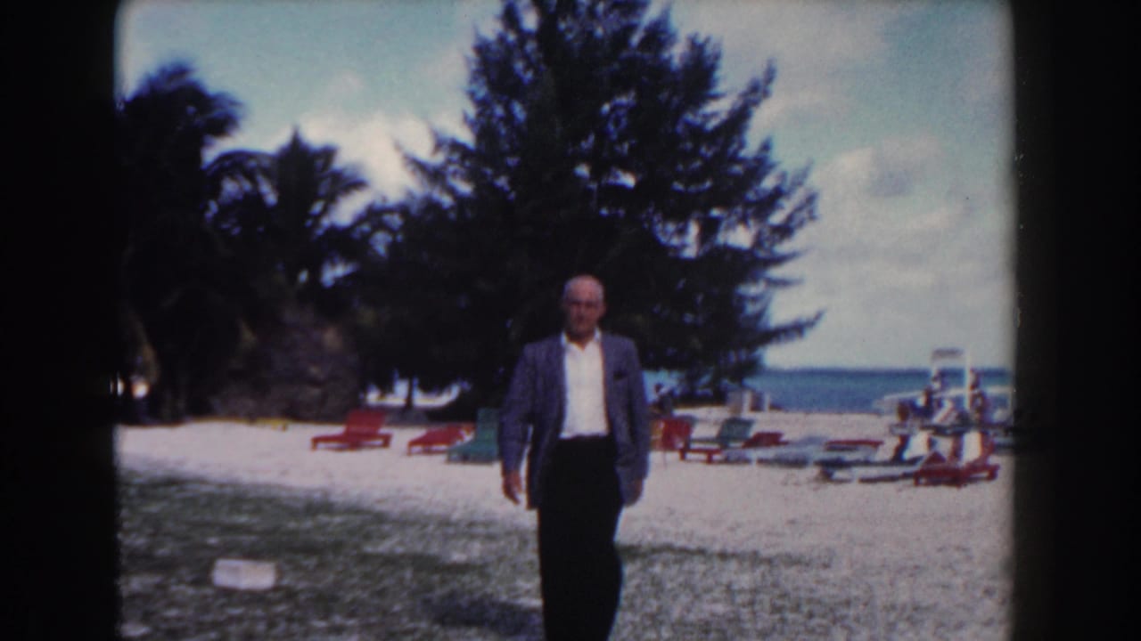 In Nassau, Bahamas in 1961, a man in a blazer walks on a beach past lounge chairs, palms, and the ocean horizon.