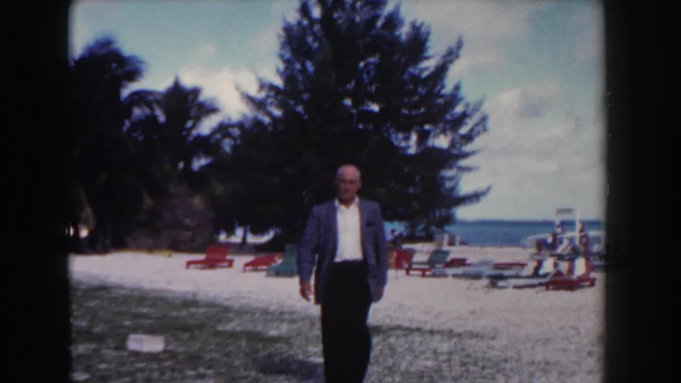In Nassau, Bahamas in 1961, a man in a blazer walks on a beach past lounge chairs, palms, and the ocean horizon.