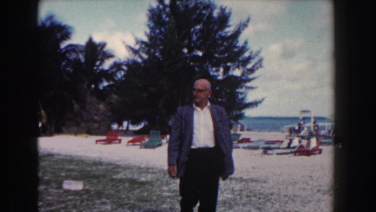 Man in a blazer standing near a beach with lounge chairs, palm trees, and water in the background.