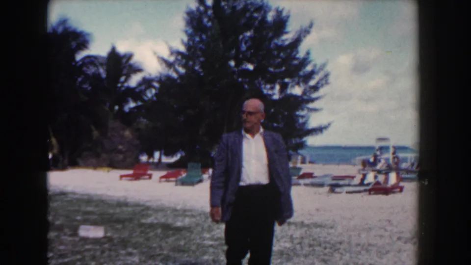 The man pauses or turns on a Nassau beach, with colorful lounge chairs, palms, and the sea visible behind him.