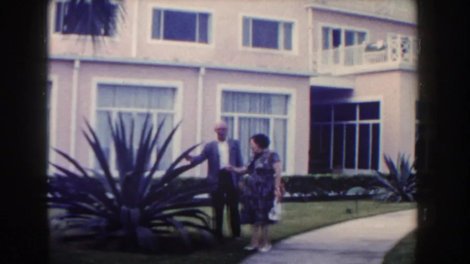 A man and woman meet outside a pink Nassau hotel building in 1961, standing near a walkway, lawn, and large agave-like plant.