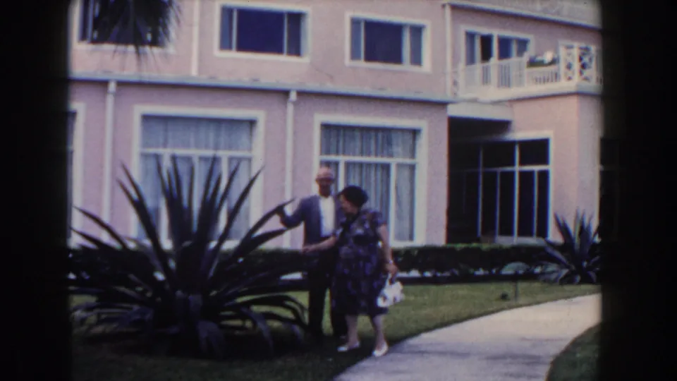 The couple walks together outside the Nassau hotel, passing a manicured lawn, walkway, windows, balcony, and tropical landscaping.