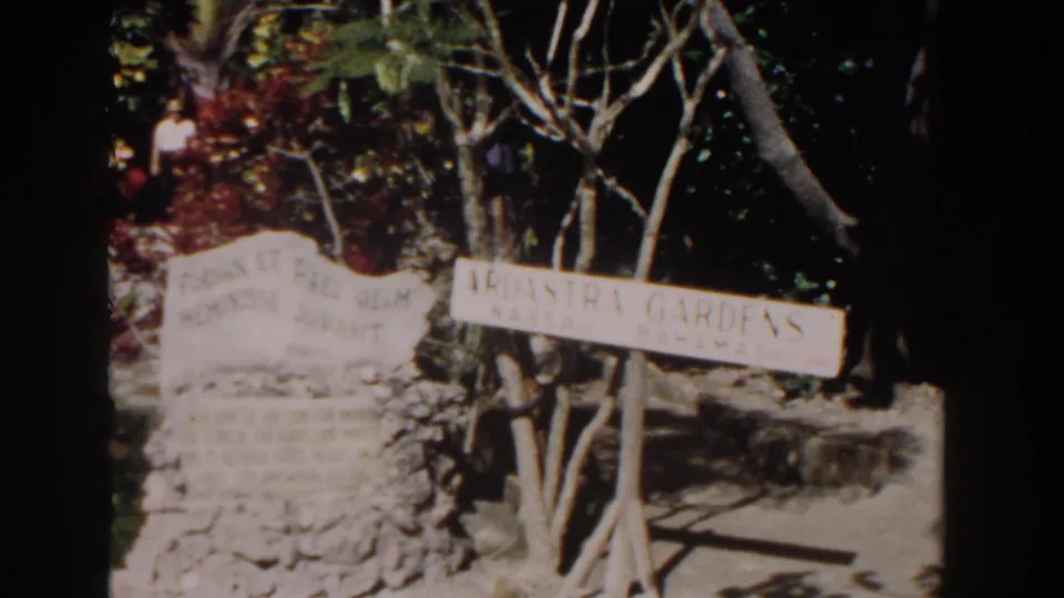 1961 Nassau frame showing the Ardastra Gardens sign, stone marker, garden path, trees, and partially visible visitors behind foliage.