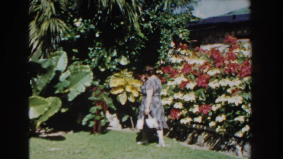 A 1961 Nassau home-movie frame shows the woman crossing the lawn near broad tropical leaves and a colorful flower border.