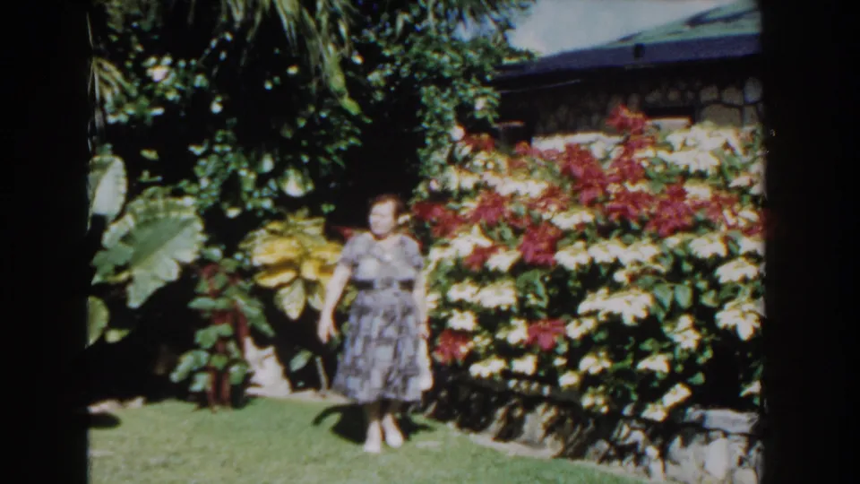 In Nassau, Bahamas in 1961, a woman in a patterned dress stands in bright sun near a tropical flower bed and stone wall.