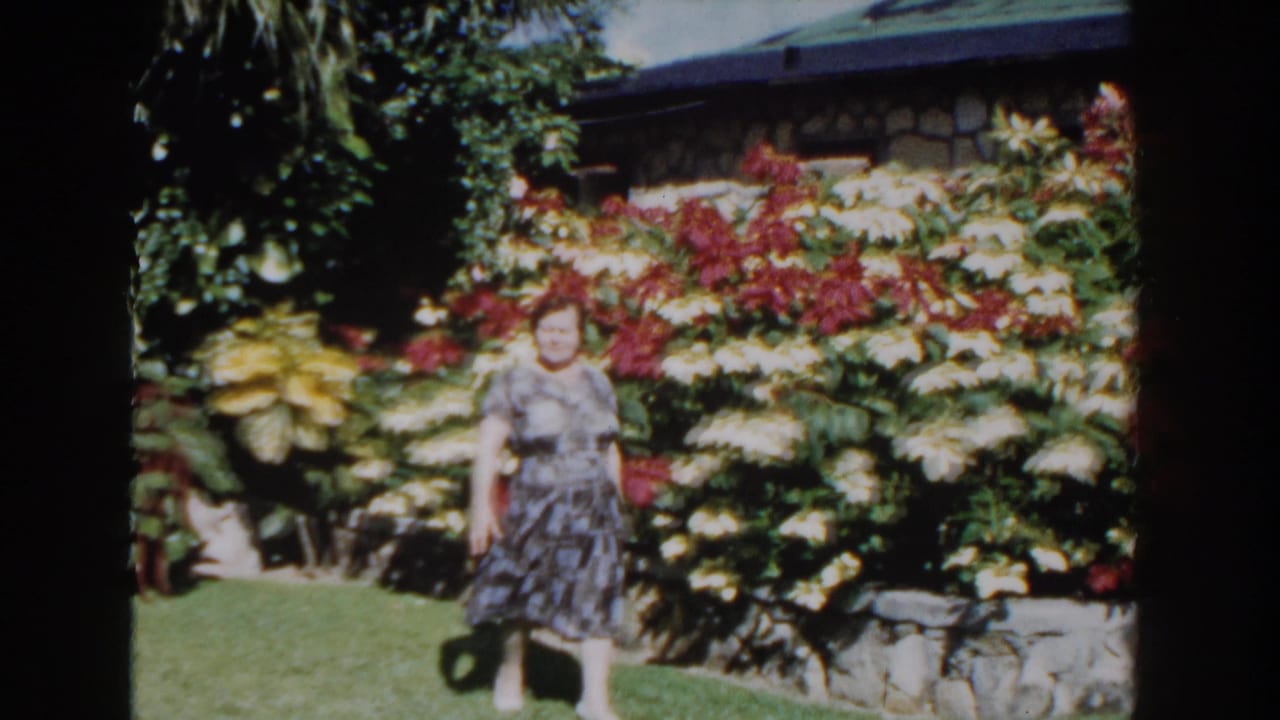 A smiling woman walks through a Nassau garden in 1961, with flowering plants, palm foliage, and a low stone wall behind her.