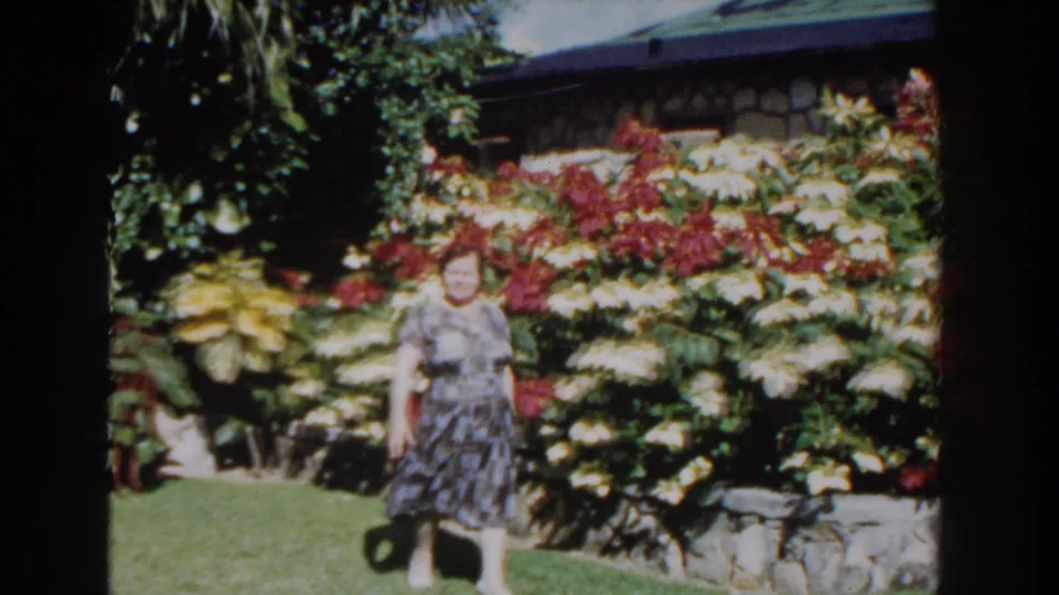 A smiling woman walks through a Nassau garden in 1961, with flowering plants, palm foliage, and a low stone wall behind her.