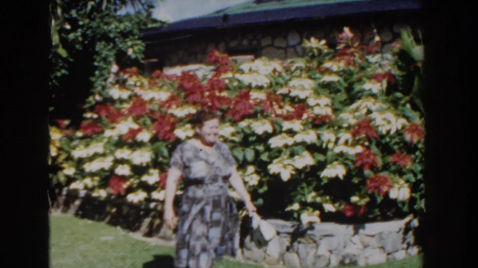 The woman walks nearer the camera holding a light purse, framed by a sunlit Nassau garden and a wall of tropical flowers.