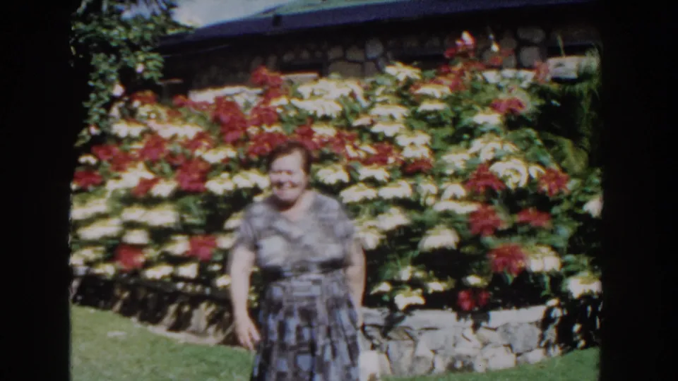 A smiling woman in a patterned dress appears closer to the camera in the 1961 Nassau garden, with dense flowers and stonework behind her.