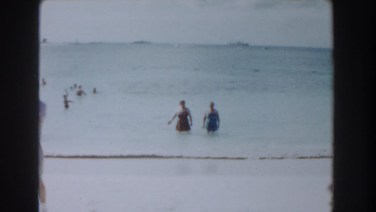 Two women wade in shallow ocean water with swimmers and a distant ship on the horizon.