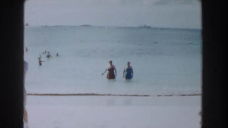 1961 Nassau beach home-movie frame showing two women wading through shallow water as other swimmers spread across the calm sea.