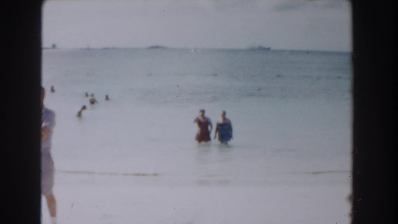 1961 Nassau beach frame with two women in shallow water, background bathers, and a partial passerby near the center frame edge.