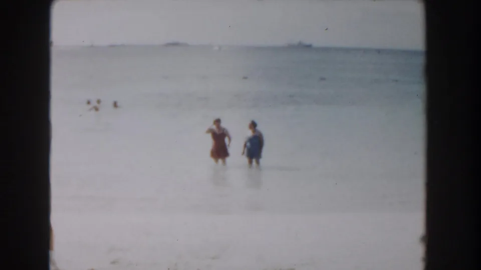 1961 Nassau beach home-movie frame showing two women wading near the shoreline beneath bright sun and soft 8mm color.