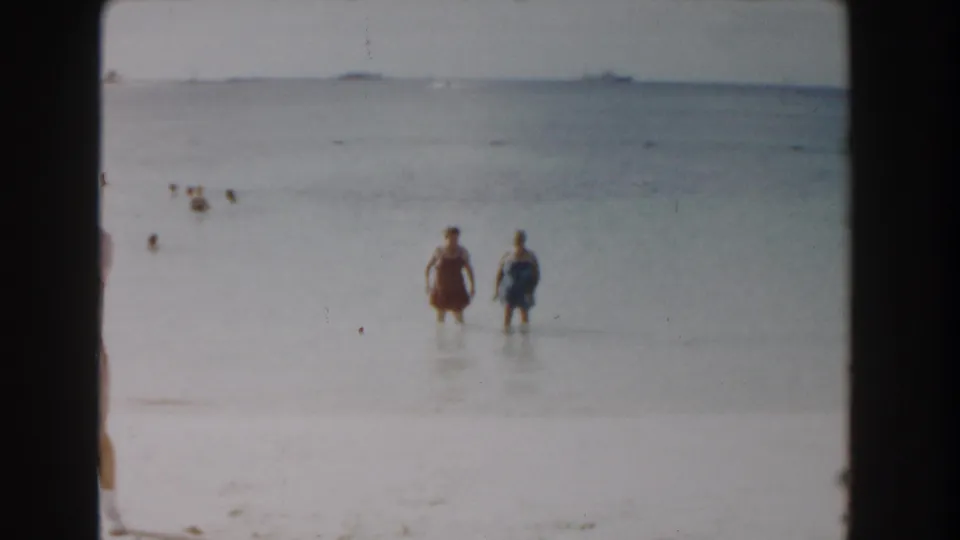 1961 Nassau beach frame with two women standing in shallow turquoise water, swimmers behind them, and ships visible along the horizon.