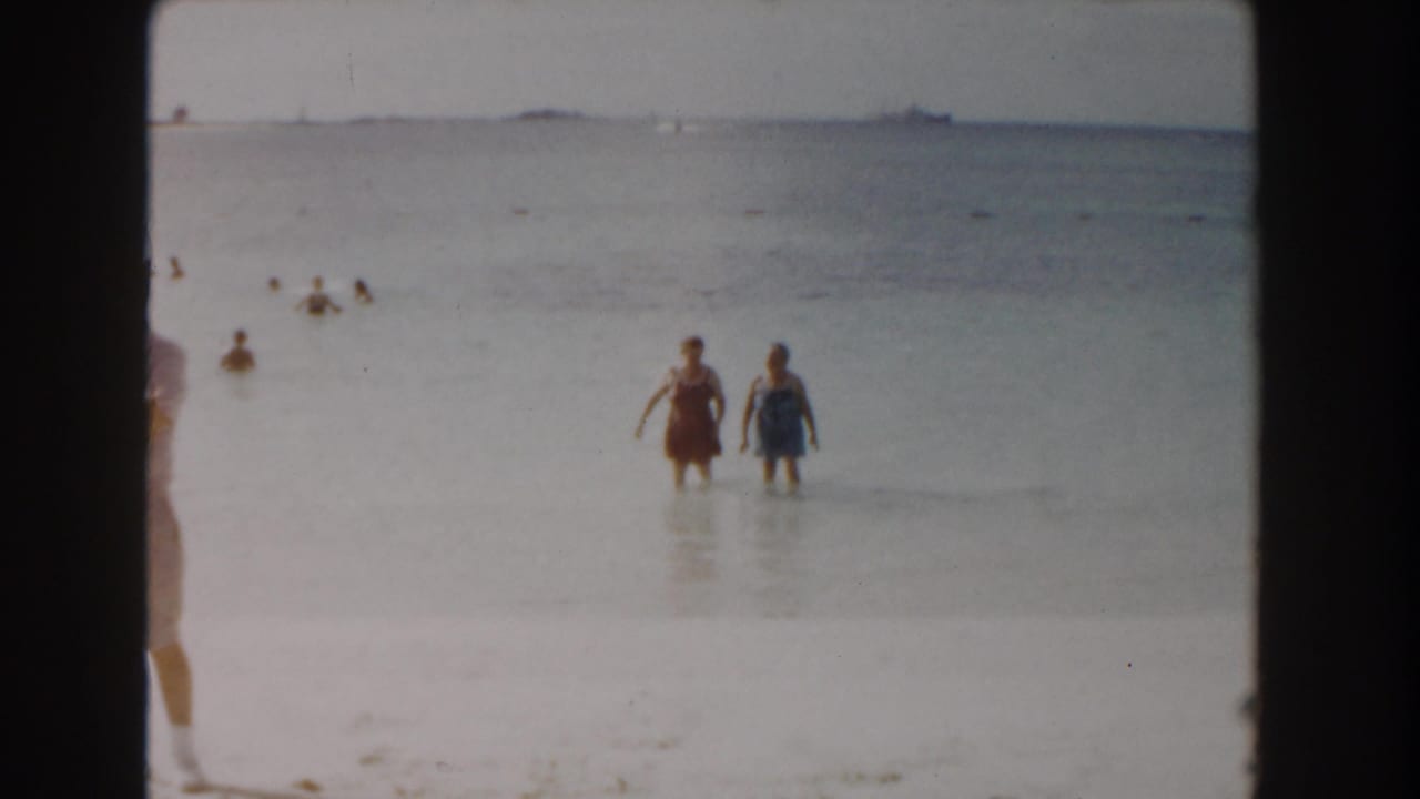 Two women wade in shallow sea near a bright sandy beach with distant bathers and boats on the horizon.