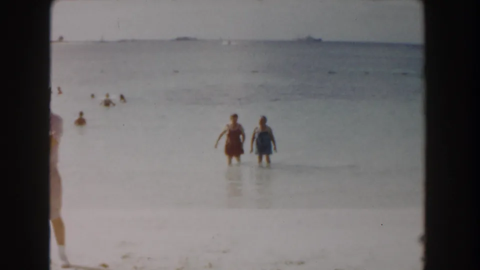 In Nassau, Bahamas, 1961, two women in swimsuits walk through shallow surf as other bathers and distant boats appear behind them.